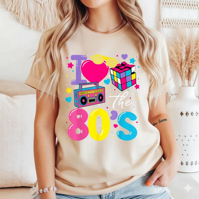 Op de afbeelding: Beige T-shirt met een kleurrijk grafisch ontwerp. Het ontwerp bevat de tekst "I love the 80's" met een roze hart, een boombox, een Rubik's Cube en andere elementen in 80's-stijl in heldere kleuren. Het shirt heeft korte mouwen en een ronde hals.