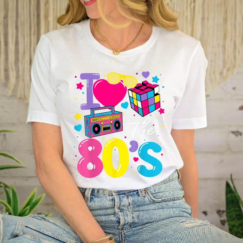 Op de afbeelding: Wit T-shirt met een kleurrijk "I Love the 80's" ontwerp. Het ontwerp bevat een roze hart, een boombox, een Rubik's kubus en andere elementen uit de jaren 80. Het T-shirt heeft een ronde hals.