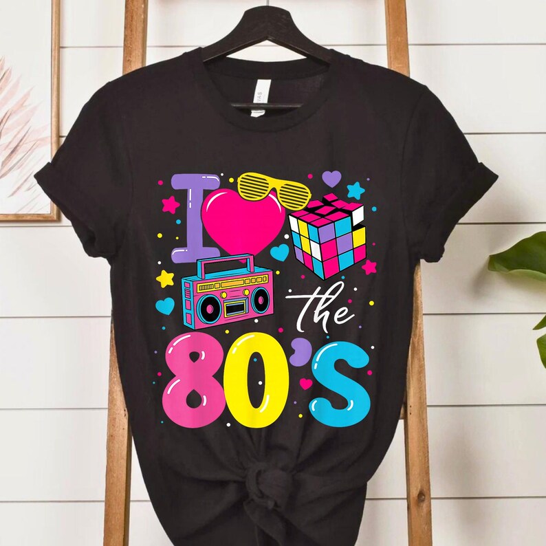 Op de afbeelding: Zwart t-shirt met een kleurrijk grafisch ontwerp. Het ontwerp bevat de tekst "I love the 80's" met een hart, zonnebril, een boombox en een Rubik's Cube. Het ontwerp is roze, geel, blauw en paars.