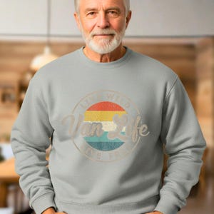 May include: A light gray sweatshirt with a circular graphic that reads "Van Life" in a vintage style. The graphic features a retro sunset design with red, yellow, and blue stripes. The text "Live Wild, Live Free" is also included.