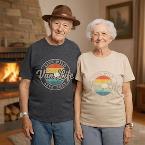 May include: Two people wearing matching t-shirts with the text "Live Wild Van Life Live Free" in a circular design with a retro color scheme. One shirt is dark gray, the other is beige. The person on the left is wearing a brown hat.