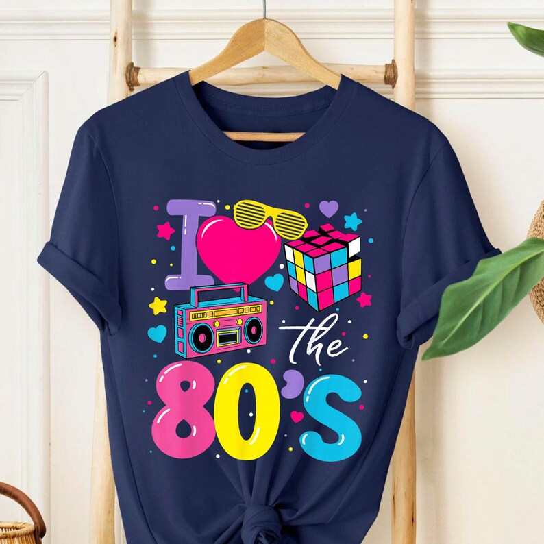 Op de afbeelding: Marineblauwe t-shirt met een kleurrijk grafisch ontwerp. Het ontwerp bevat de woorden "I love the 80's" met een hart, zonnebril, Rubik's Cube en een boombox. Het t-shirt hangt aan een houten hanger.