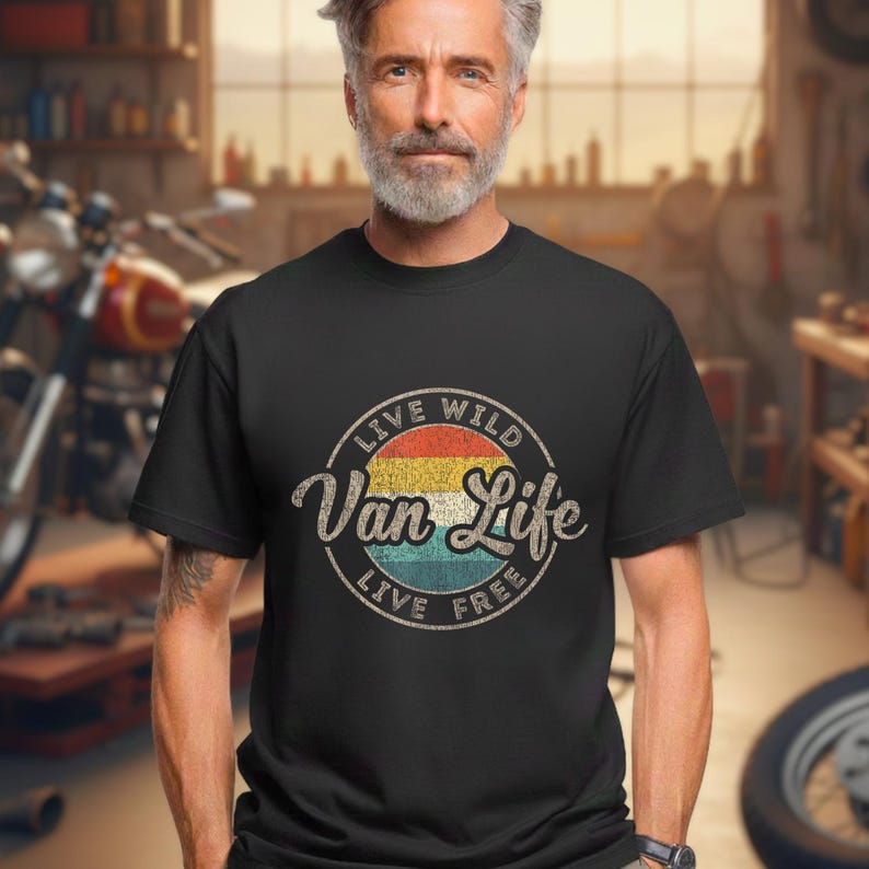 May include: Black t-shirt featuring a circular graphic with the text "Live Wild Van Life Live Free." The graphic has a retro design with a color gradient of red, yellow, and blue. The t-shirt is worn by a person with a gray beard.