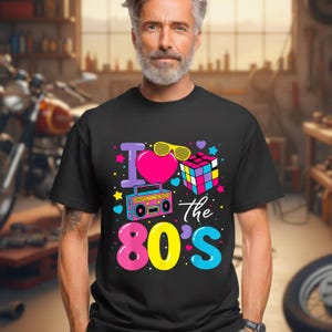 Op de afbeelding: Zwart T-shirt met een kleurrijk grafisch ontwerp. Het ontwerp bevat de tekst "I ❤️ the 80's" met een hart, zonnebril, Rubik's Cube en een boombox. Het T-shirt heeft een ronde hals.