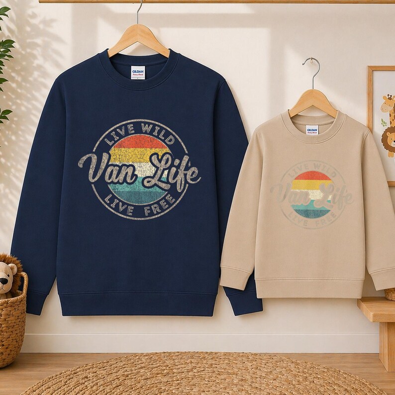 May include: Two crewneck sweatshirts, one navy blue and one tan, hang on wooden hangers. Both feature a circular graphic with the text "Live Wild Van Life Live Free" in a vintage style. The graphic includes a sunset color scheme.