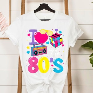 Op de afbeelding: Wit T-shirt met een kleurrijk grafisch ontwerp. Het ontwerp bevat de tekst "I love the 80's" met een hart, zonnebril, Rubik's kubus en een boombox. De afbeelding is roze, geel, blauw, paars en groen.