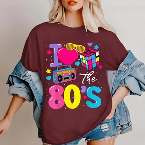Op de afbeelding: Bordeaux t-shirt met een kleurrijk jaren 80 thema. Het ontwerp bevat een roze hart, gele zonnebril, een Rubik's Cube en een boombox. De tekst "I love the 80's" is ook aanwezig.