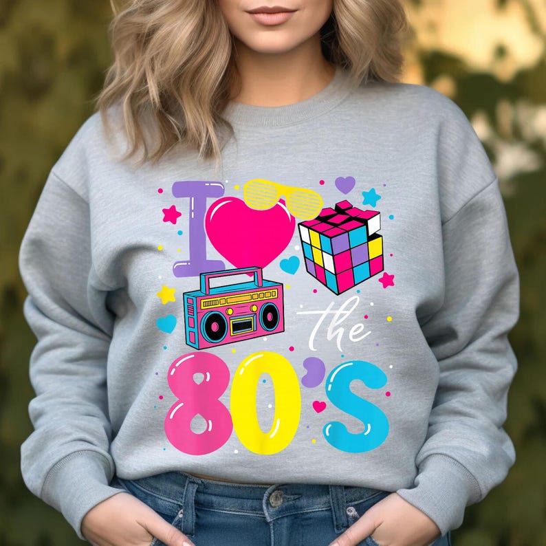 Op de afbeelding: Een grijze sweatshirt met een kleurrijke afbeelding met de tekst "I love the 80s". Het ontwerp bevat een roze hart, een Rubik's Cube, een boombox en andere elementen met een jaren 80-thema.