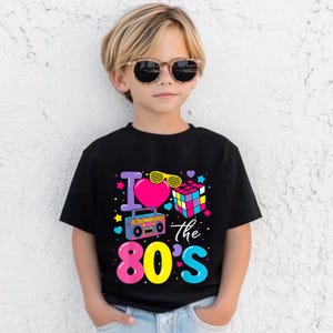 Op de afbeelding: Zwart T-shirt met een kleurrijke afbeelding met de tekst "I love the 80's". De afbeelding bevat een roze hart, gele zonnebril, een Rubik's kubus en een boombox. Het T-shirt wordt gedragen door een kind met een zonnebril.