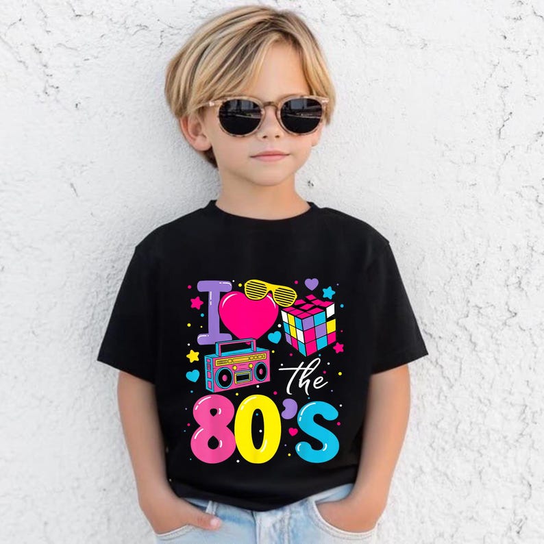 Op de afbeelding: Zwart T-shirt met een kleurrijke afbeelding met de tekst "I love the 80's". De afbeelding bevat een roze hart, gele zonnebril, een Rubik's kubus en een boombox. Het T-shirt wordt gedragen door een kind met een zonnebril.