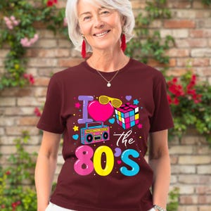 Op de afbeelding: Een bordeauxrood t-shirt met een kleurrijke afbeelding met de tekst "I love the 80's". De afbeelding bevat een roze hart, een Rubik's kubus, een boombox en gele zonnebrillen. Het t-shirt wordt door een persoon gedragen.