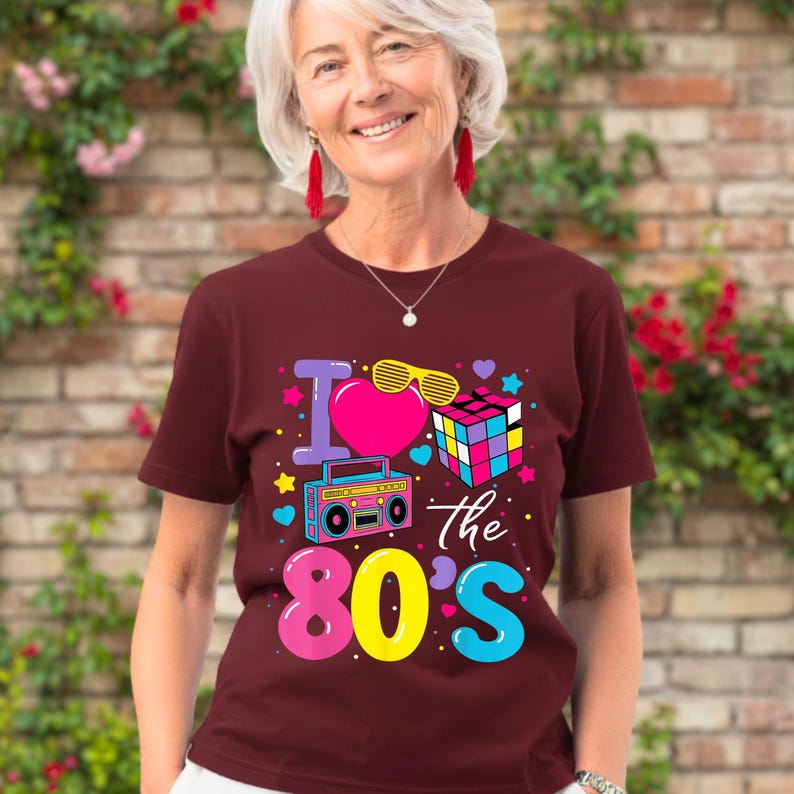 Op de afbeelding: Een bordeauxrood t-shirt met een kleurrijke afbeelding met de tekst "I love the 80's". De afbeelding bevat een roze hart, een Rubik's kubus, een boombox en gele zonnebrillen. Het t-shirt wordt door een persoon gedragen.
