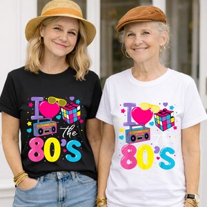 Op de afbeelding: Twee vrouwen dragen T-shirts met een kleurrijk "I Love the 80's" ontwerp. Het ontwerp bevat een roze hart, zonnebril, Rubik's kubus en een boombox. E&eacute;n shirt is zwart, de andere is wit.