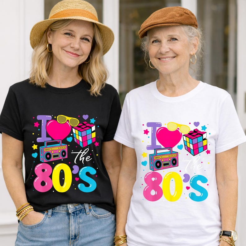 Op de afbeelding: Twee vrouwen dragen T-shirts met een kleurrijk "I Love the 80's" ontwerp. Het ontwerp bevat een roze hart, zonnebril, Rubik's kubus en een boombox. E&eacute;n shirt is zwart, de andere is wit.