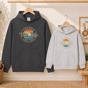 May include: Two hooded sweatshirts, one charcoal gray and one light gray, each with a front pocket and a hood. The charcoal gray sweatshirt features a circular graphic with the text "Live Wild Van Life Live Free". The light gray sweatshirt has the same graphic.