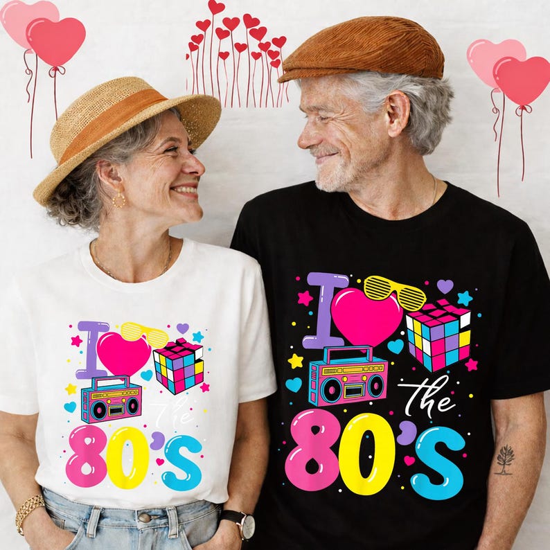 Op de afbeelding: Een stel draagt bijpassende t-shirts met een retro jaren 80 design. De shirts hebben een roze hart, zonnebril, Rubik's kubus en een boombox. De tekst op de shirts luidt "I love the 80's" in levendige kleuren.