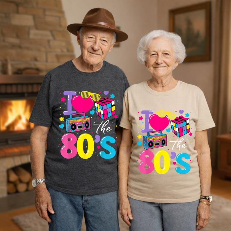 Op de afbeelding: Twee mensen dragen bijpassende t-shirts met een kleurrijk "I Love the 80's" ontwerp. Het ontwerp bevat een roze hart, zonnebril, Rubik's kubus en een boombox. De shirts zijn donkergrijs en beige.