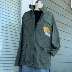 May include: An olive green utility jacket with a high collar, button-down front, and two chest pockets. A yellow and white patterned heart patch is stitched onto the left chest. The jacket is layered over a black top and a blue denim mini skirt.
