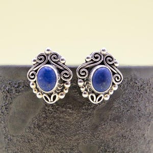 Puede incluir: Un par de pendientes de botón plateados con piedras ovaladas de lapislázuli azul oscuro. Los pendientes tienen un diseño ornamental de filigrana con detalles en espiral y pequeños acentos de cuentas.