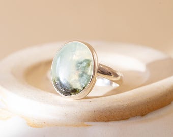 May include: A silver ring featuring a smooth, oval-shaped cabochon stone. The stone is translucent with pale green and dark mossy inclusions visible throughout its surface. The metal band is polished and simple, holding the stone in a bezel setting.