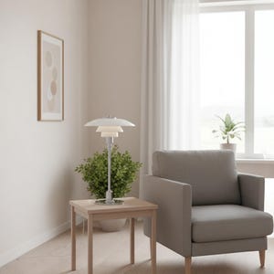 Peut inclure: Une lampe de table blanche et argentée avec un abat-jour à gradins est posée sur une petite table en bois. La lampe est à côté d'une petite plante verte et d'un fauteuil gris. Une œuvre d'art abstraite encadrée est accrochée au mur.