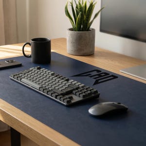 Puede incluir: Una configuración de escritorio moderna con una alfombrilla azul marino con la palabra "CAD" impresa en negro. Un teclado y un ratón plateados están sobre la alfombrilla, junto a una taza negra, un smartphone y una planta en maceta.