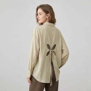 Könnte beinhalten: Beige Langarmhemd mit einzigartigem offenem Rückendesign. Der Rücken hat einen schmetterlingsförmigen Ausschnitt mit Bindebändern. Das Hemd hat einen klassischen Kragen und geknöpfte Manschetten. Der Stoff scheint ein leichtes, strukturiertes Material zu sein.