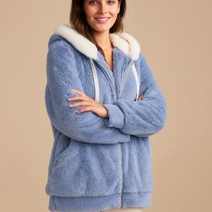 May include: A light blue, hooded zip-up jacket with a fluffy, textured appearance. The hood is lined with a cream-colored material, and the jacket has a full-length zipper and white drawstrings. The jacket has two front pockets and is made of a soft, plush fabric.