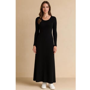 以下が含まれることがあります： 長袖と丸いネックラインの黒いロングドレス。 ドレスはリブ編みのテクスチャで、白いスニーカーを履いています。 女性はベージュの背景の前に立っています。