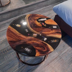 Puede incluir: Mesa de centro redonda con tapa de resina negra con vetas de madera e incrustaciones de botellas de licor en miniatura. La mesa tiene una base de metal color bronce y está sobre un suelo de madera gris.