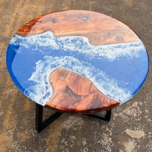 Op de afbeelding: Ronde salontafel met een uniek ontwerp. Het tafelblad combineert hout en blauwe hars, wat een oceaanscène creëert. Het staat op een zwart metalen onderstel. Het hout heeft een roodbruine kleur. De diameter is ongeveer 90 cm.