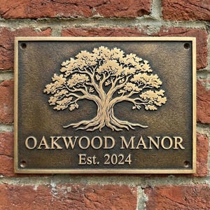 Op de afbeelding: Een rechthoekige bronzen plaat met de woorden "OAKWOOD MANOR" en "Est. 2024" in een schreeflettertype. Een gedetailleerd goudkleurig boomontwerp is gecentreerd boven de tekst. De plaat is bevestigd aan een rode bakstenen muur.