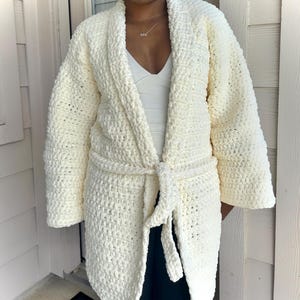 May include: Cream-colored, open-front crochet robe with a matching belt. The robe features a thick, textured stitch and long sleeves. The belt is tied at the waist. This cozy robe is perfect for lounging.