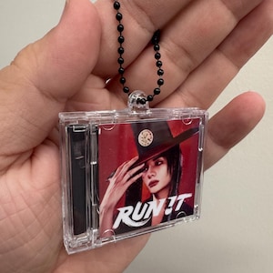 Peut inclure: Un porte-clés miniature en forme de boîtier CD en plastique transparent avec une chaînette à billes noire. La pochette montre une personne portant un chapeau à larges bords avec un emblème circulaire sur fond rouge et le texte blanc RUN IT.