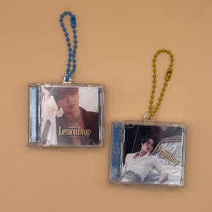 May include: Two clear plastic CD-shaped keyrings. Each keyring features a photo of a person and the text "Lemon Drop". One keyring has a blue chain, the other has a gold chain. The background is a solid tan colour.
