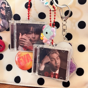 May include: A cream-coloured bag with black polka dots decorated with two clear plastic miniature CD case keychains featuring portraits of men, a safety pin with beads spelling BTS, a small cartoon character charm, and circular pinback buttons.