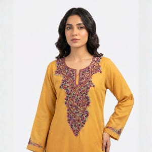 Puede incluir: Túnica de color amarillo mostaza con mangas largas y bordados florales intrincados en tonos morados, verdes y rosas alrededor del cuello y los puños. La túnica tiene un diseño simple y elegante.