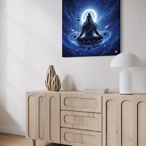 Può includere: Una stampa artistica su tela quadrata raffigura una figura meditante in posizione del loto, su uno sfondo blu cosmico con un alone luminoso. La figura tiene un tridente. L'opera d'arte è esposta sopra un mobile in legno chiaro con vasi decorativi e una lampada moderna.