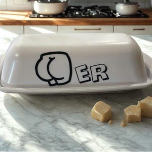 Op de afbeelding: Een witte keramische boterdoos met een deksel met een zwarte afbeelding van een kont en de letters "ER". Verschillende blokjes boter liggen op het marmeren oppervlak. Keukengerei.