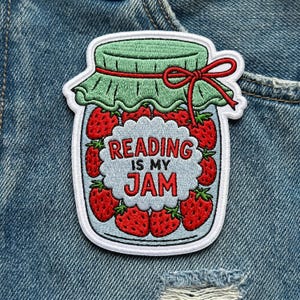 May include: Embroidered patch featuring a jar filled with strawberries. The jar has a green lid and a red bow. The text "READING IS MY JAM" is in the center. The patch is on a denim background.