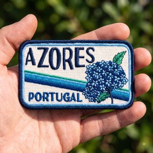 May include: A rectangular embroidered patch with a cream background, featuring the dark blue text AZORES and PORTUGAL. A diagonal blue and green stripe crosses the center, and a cluster of blue hydrangea flowers with green leaves is on the right.