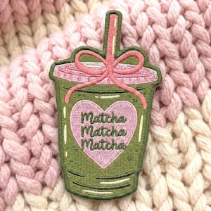 May include: An embroidered patch shaped like a green matcha drink in a cup with a lid and straw. A pink bow sits atop the lid, and a large pink heart in the center features the repeated text Matcha, Matcha, Matcha in dark green embroidery.