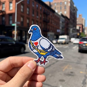May include: An embroidered iron-on patch shaped like a pigeon with blue and white feathers, red feet, and a red eye. The pigeon wears a yellow fanny pack and a white chest strap featuring a red heart icon and the blue text NY.
