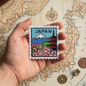 May include: Embroidered patch featuring a Japanese landscape with Mount Fuji, a pagoda, and cherry blossoms. The patch is in the shape of a postage stamp with the word "JAPAN" at the top. The patch is held in a hand.