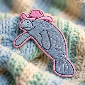May include: An embroidered patch featuring a grey manatee wearing a pink cowboy hat. The manatee has a rounded snout, a curved body, and a flat tail, all outlined in a thick pink border with white stitch detailing on its body and tail.