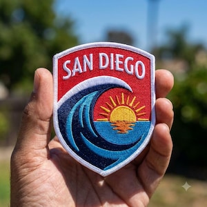 May include: A shield-shaped embroidered patch featuring a red background with the white text SAN DIEGO at the top. Below the text is a large dark blue and light blue ocean wave crashing over a yellow sun rising above a light blue horizon.
