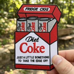 May include: Embroidered patch featuring a red and white Diet Coke cigarette pack design. The patch reads "FRIDGE CIGS" at the top and "Diet Coke" in the center, with the phrase "JUST A LITTLE SOMETHING TO TAKE THE EDGE OFF" below.