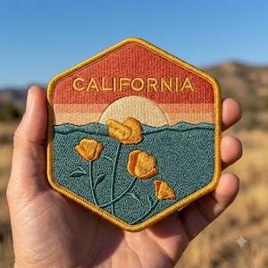 May include: A hexagonal embroidered patch featuring a sunset landscape with the word CALIFORNIA stitched in gold thread at the top. The design includes a cream sun, a dark teal foreground with four yellow poppy flowers, and a red-orange sky.