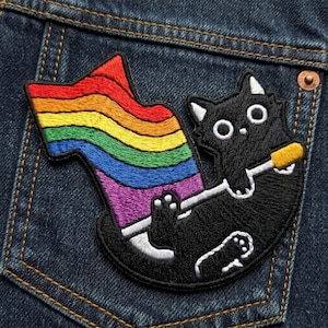 May include: Embroidered patch featuring a black cat holding a rainbow flag. The cat has white accents and large, round eyes. The patch is sewn onto a blue denim pocket.