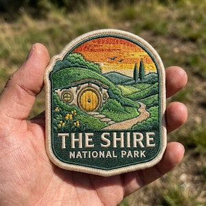 May include: An embroidered patch featuring a landscape with a round yellow door set into a green hill, a winding path, and a sunset sky. The text THE SHIRE NATIONAL PARK is stitched in white at the bottom against a dark green background.